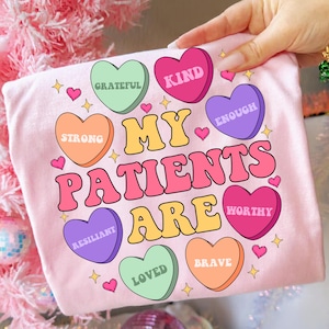 Może przedstawiać: Jasnoróżowa koszulka z napisem "MY PATIENTS ARE" dużymi, kolorowymi literami. Wokół tekstu znajdują się cukierki w kształcie serca ze słowami takimi jak "KIND" i "WORTHY". Małe serca i gwiazdki rozrzucone są po całym projekcie.