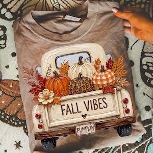 May include: A tan t-shirt featuring a graphic of a vintage truck carrying pumpkins and autumn leaves. The truck bed displays the text FALL VIBES and a small heart. The pumpkins include a solid orange one, a white one, and a leopard print one.