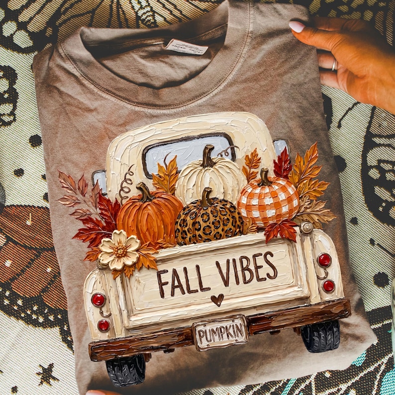 May include: A tan t-shirt featuring a graphic of a vintage truck carrying pumpkins and autumn leaves. The truck bed displays the text FALL VIBES and a small heart. The pumpkins include a solid orange one, a white one, and a leopard print one.