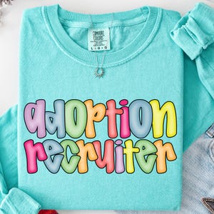 May include: A turquoise Comfort Colors t-shirt with the words "Adoption Recruiter" in colorful bubble letters. The shirt has a crew neck and a small tag at the neckline. A silver necklace with a sunburst pendant hangs on the shirt.