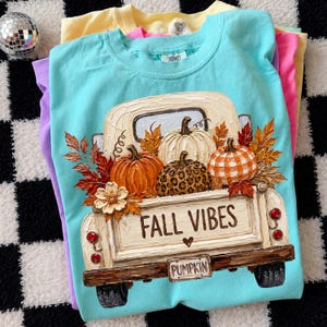 May include: A turquoise t-shirt featuring a graphic of a cream-colored vintage truck carrying three pumpkins, one orange, one white, and one with a leopard print. The truck bed displays the text FALL VIBES and a license plate reading PUMPKIN.