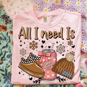 May include: Pink Comfort Colors t-shirt with the text "All I Need Is" and graphics of a Dr. Pepper can, brown shoes, and a knit hat. The Dr. Pepper can has a pink bow and a straw. The hat has a checkered pattern.