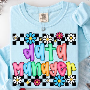 May include: Light blue long-sleeve shirt with the words "data manager" in colorful bubble letters. The shirt features a black and white checkered pattern with daisy and flower graphics. A silver necklace is visible.