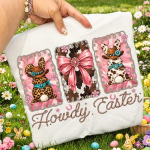 May include: White t-shirt featuring three framed designs with a pink floral border. The designs include a cowhide bow, and two bunnies wearing cowboy hats and bandanas. The text "Howdy Easter" is written in a rope-like font.