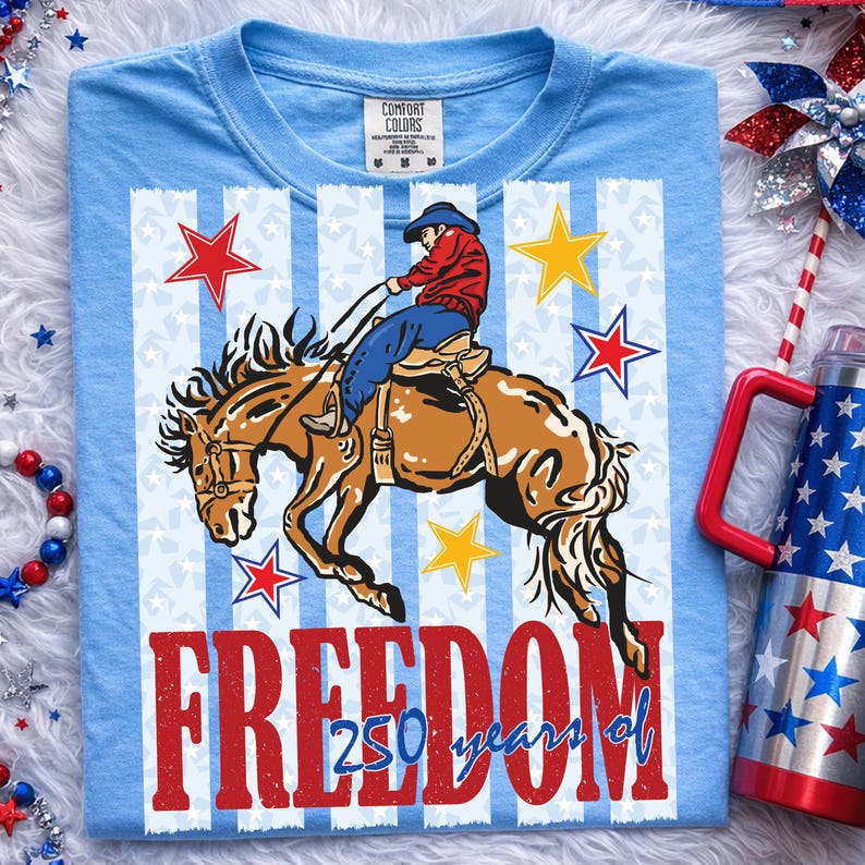 May include: A light blue t-shirt featuring a graphic of a cowboy riding a bucking horse. The design includes vertical white stripes, red and yellow stars, and the red text "FREEDOM" above the blue script text "250 years of". The shirt has a Comfort Colors label.