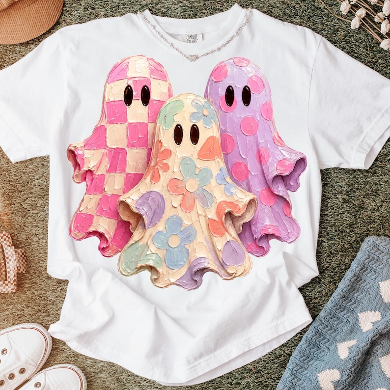 Pu&ograve; includere: Una t-shirt bianca a maniche corte con una grafica raffigurante tre fantasmi con dettagli materici simili alla pittura. Il fantasma a sinistra ha un motivo a scacchi rosa e crema, quello centrale un disegno floreale e quello a destra pois viola.