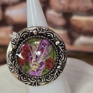 May include: A silver-toned ring with an ornate, filigree design. The ring's centerpiece features a clear resin dome encasing a small animal skull, red and purple roses, and green leaves. The ring is displayed on a white stand.