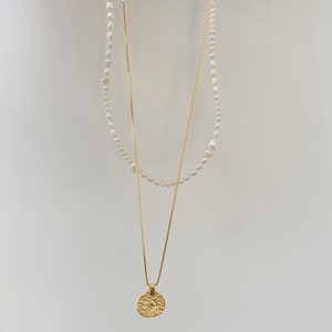 May include: A gold chain necklace with a textured round pendant and a pearl necklace layered on top.