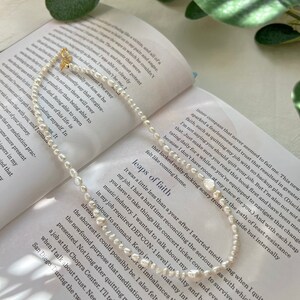 May include: A delicate pearl necklace with a gold clasp, resting on an open book with text about taking leaps of faith. The book's pages are visible in the background.