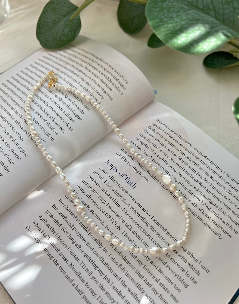 May include: A delicate pearl necklace with a gold clasp, resting on an open book with text about taking leaps of faith. The book's pages are visible in the background.