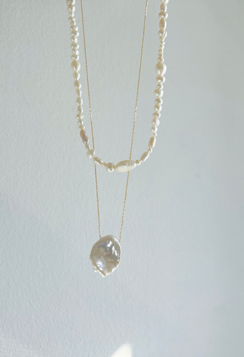 May include: A layered pearl necklace with a large, irregular pearl pendant. The necklace is made of gold chain and features white pearls of varying sizes.