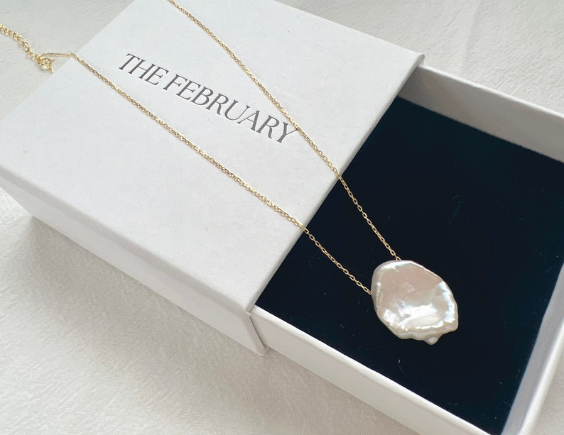 May include: A white baroque pearl pendant necklace on a gold chain in a white box with the text "THE FEBRUARY" on the lid.