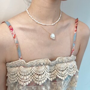 May include: A woman wearing a white pearl necklace and a gold chain with a large pearl pendant. She is wearing a light blue and white floral patterned top with a lace trim.