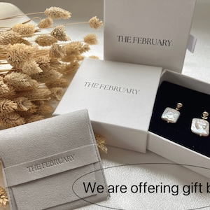 May include: Three white boxes with the text "THE FEBRUARY" printed on them. The boxes are for jewelry and are shown with a pair of pearl earrings inside one of the boxes. The boxes are on a white surface with dried flowers in the background.