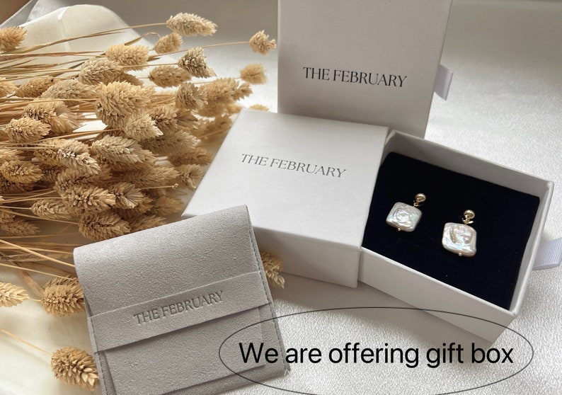 May include: Three white boxes with the text "THE FEBRUARY" printed on them. The boxes are for jewelry and are shown with a pair of pearl earrings inside one of the boxes. The boxes are on a white surface with dried flowers in the background.