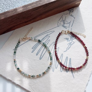 May include: Two delicate bracelets with gold accents. One bracelet features green and white agate beads, while the other has deep red garnet beads.