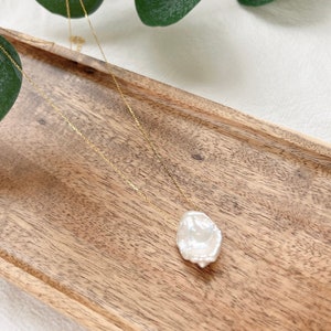 May include: A delicate gold chain necklace with a large, irregular-shaped pearl pendant. The pearl is white and has a slightly textured surface. The necklace is displayed on a wooden tray.
