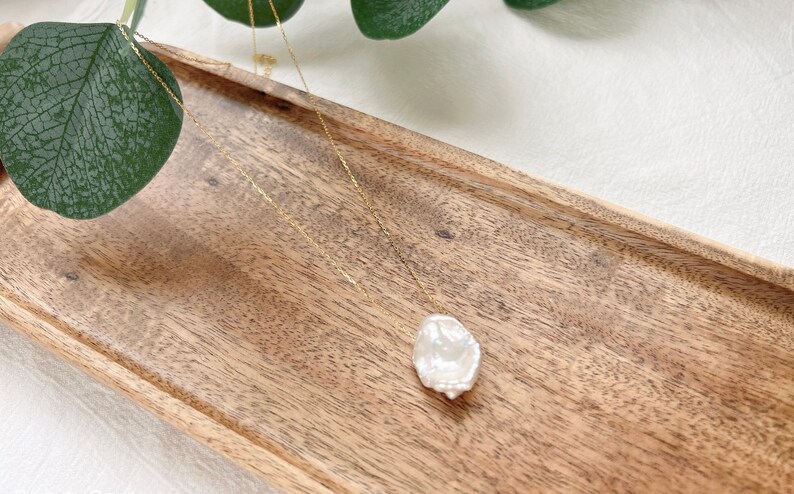 May include: A delicate gold chain necklace with a large, irregular-shaped pearl pendant. The pearl is white and has a slightly textured surface. The necklace is displayed on a wooden tray.
