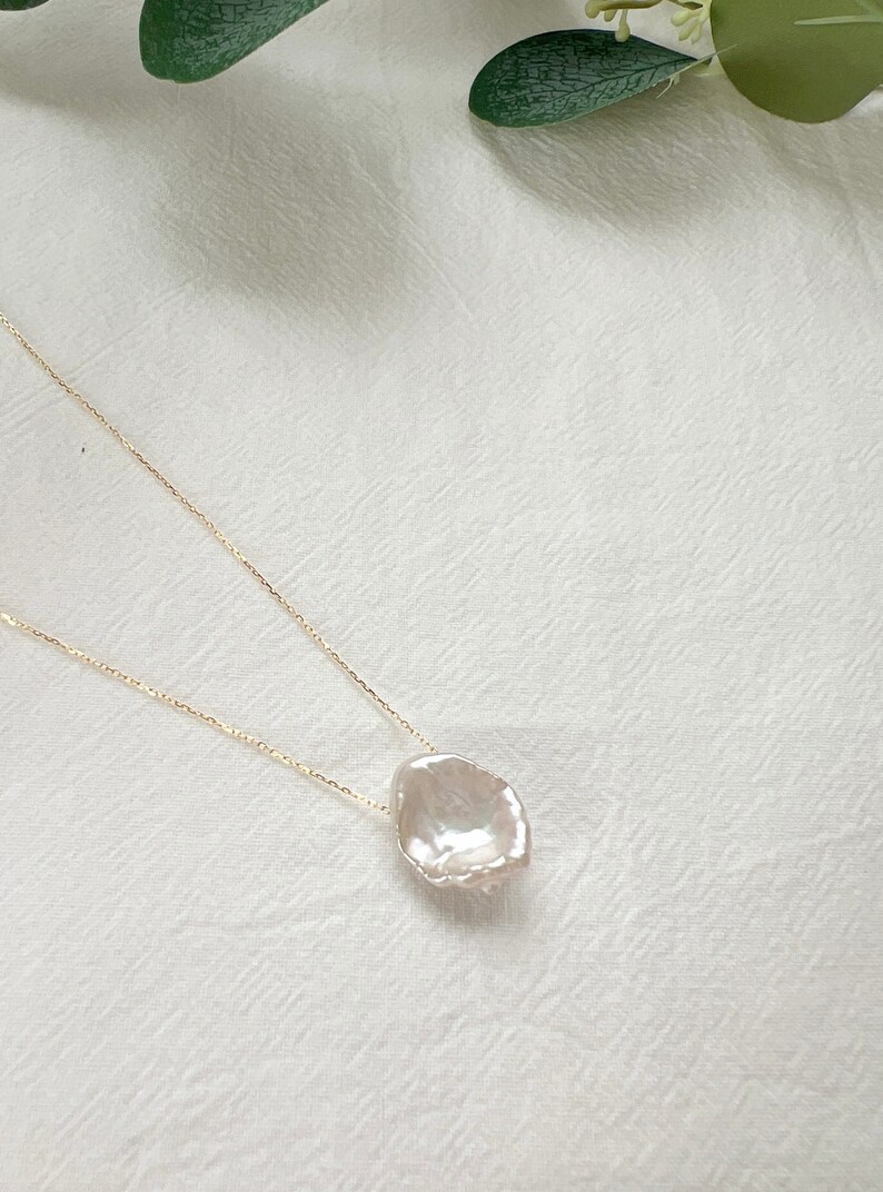 May include: A delicate gold chain necklace with a large, irregular-shaped pearl pendant. The pearl is white and has a slightly iridescent sheen.