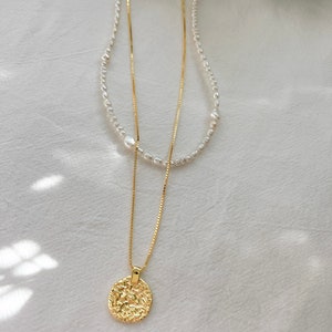 May include: A gold chain necklace with a textured round pendant and a pearl necklace layered on top.