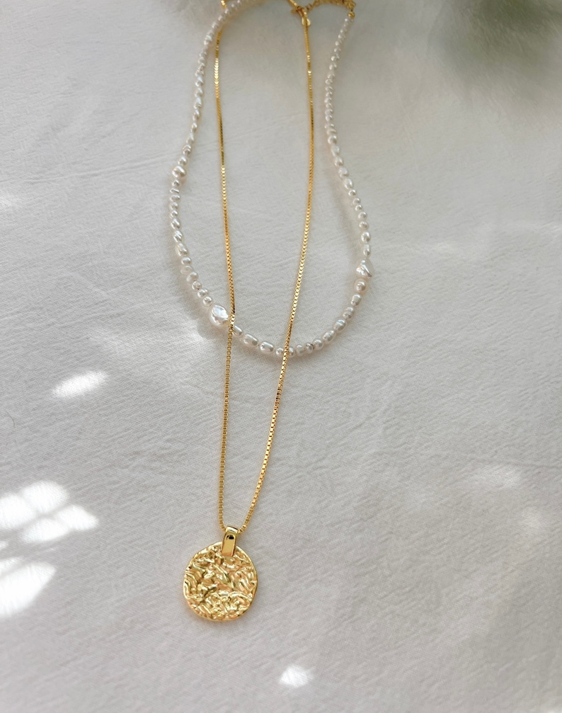 May include: A gold chain necklace with a textured round pendant and a pearl necklace layered on top.