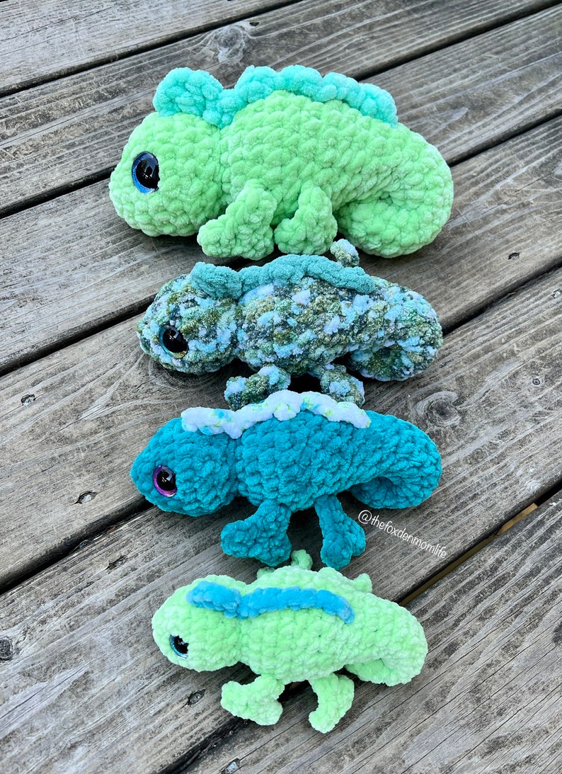 May include: Four crocheted chameleon plushies in different sizes and colours. The largest is green, the next is green and white, the next is teal, and the smallest is green with a blue stripe.  The plushies are on a wooden surface.  @thefoxdenmomlife