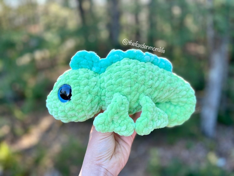 May include: A green crocheted chameleon plush toy with large blue eyes. The toy is being held in a hand with a blurred green background.
