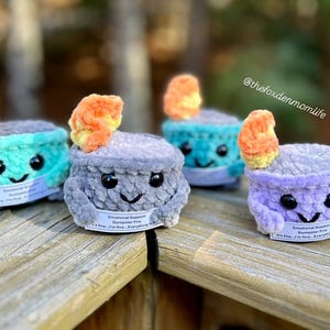 May include: Four crocheted plush toys shaped like a campfire with a flame on top. Each toy has a different color and a smiling face. The toys have a tag that reads "Emotional Support Dumpster Fire" and "It's fine, I'm fine, Everything is fine."