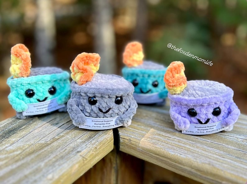 May include: Four crocheted plush toys shaped like a campfire with a flame on top. Each toy has a different color and a smiling face. The toys have a tag that reads "Emotional Support Dumpster Fire" and "It's fine, I'm fine, Everything is fine."