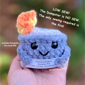Könnte beinhalten: Ein gehäkeltes Plüschtier in Form eines Mülltonnenfeuers mit einem lächelnden Gesicht. Das Spielzeug ist blau und grau mit einer leuchtend orangefarbenen und gelben Flamme oben drauf. Das Spielzeug hat ein Etikett mit der Aufschrift "Emotional Support Dumpster Fire it's fine...I'm fine...Everything is fine..." und "@thefoxdenmomlife".