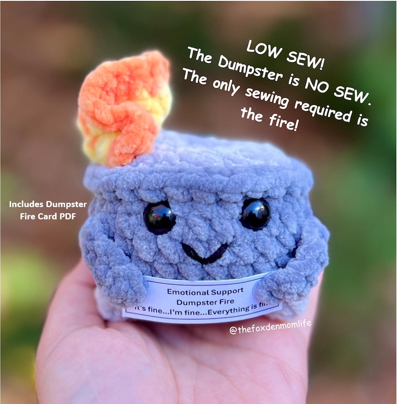 May include: A crocheted plush toy shaped like a dumpster fire with a smiling face. The toy is blue and gray with a bright orange and yellow flame on top. The toy has a tag that reads "Emotional Support Dumpster Fire it's fine...I'm fine...Everything is fine..." and "@thefoxdenmomlife".