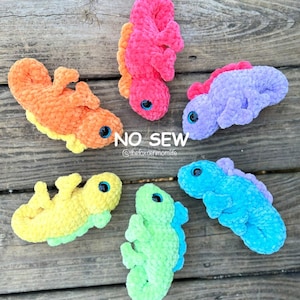 以下が含まれることがあります： ピンク、オレンジ、パープル、ブルー、グリーンの5色の編みぐるみトカゲ。各トカゲは黒い目を2つ持ち、柔らかくふわふわの糸で作られています。トカゲは木製の表面に並べてあります。画像には「NO SEW」と「@thefoxdenmomlife」のテキストが表示されています。