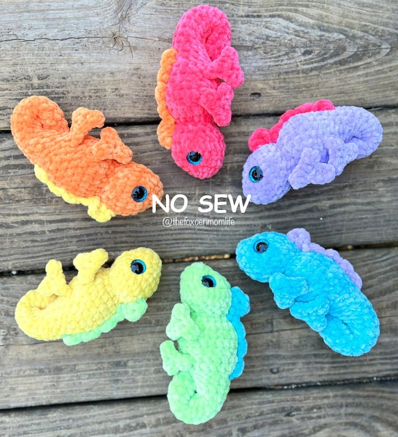 May include: Five crocheted amigurumi lizards in different colours: pink, orange, purple, blue, and green. Each lizard has a pair of black eyes and is made with a soft, fuzzy yarn. The lizards are arranged on a wooden surface. The text "NO SEW" and "@thefoxdenmomlife" is visible in the image.