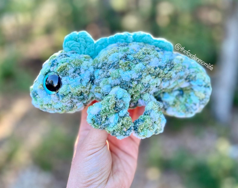 May include: A crocheted chameleon plush toy made with green, blue and white yarn. The chameleon has large, black eyes with blue pupils and a textured, bumpy surface.