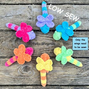 Op de afbeelding: Zes kleurrijke gehaakte libellen speelgoed met een low-sew ontwerp. De vleugels zijn los en moeten worden vastgenaaid. De libellenlichamen zijn gehaakt in verschillende kleuren, waaronder roze, blauw, groen, geel en oranje. De tekst "LOW SEW" is in wit op een blauwe achtergrond geschreven. De tekst "Only the wings need to be sewn" is in wit op een blauwe achtergrond geschreven.