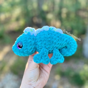 May include: A teal blue crocheted chameleon plush toy with a white and green patterned back. The toy has large purple eyes and a small pink nose.