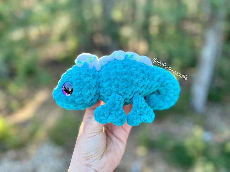 May include: A teal blue crocheted chameleon plush toy with a white and green patterned back. The toy has large purple eyes and a small pink nose.