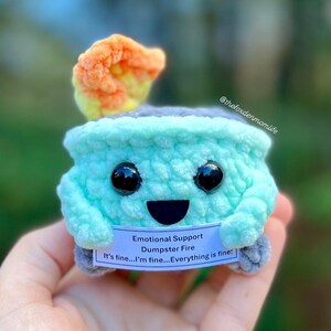 May include: A crocheted plush toy shaped like a small, green, round container with a gray lid. The toy has a smiling face with black eyes and a black mouth. The toy has a small, orange and yellow flame on top of the lid. The toy is holding a white sign that says "Emotional Support Dumpster Fire It's fine...I'm fine...Everything is fine!"