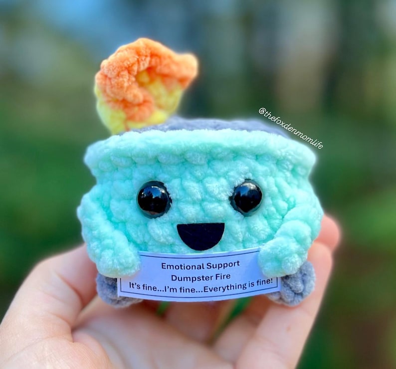 May include: A crocheted plush toy shaped like a small, green, round container with a gray lid. The toy has a smiling face with black eyes and a black mouth. The toy has a small, orange and yellow flame on top of the lid. The toy is holding a white sign that says "Emotional Support Dumpster Fire It's fine...I'm fine...Everything is fine!"
