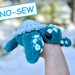May include: A teal crocheted turtle with white flower accents and large black eyes. The turtle is being held in a hand and the text "NO-SEW" is in the background. The text "@thefoxdenmomlife" is also visible in the image.