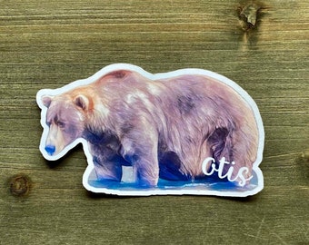 May include: A sticker featuring a watercolor illustration of a brown bear standing in water. The bear is looking to the left and the text "Otis" is written below the bear.
