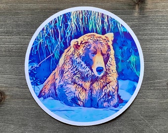 Puede incluir: Un oso marrón con un fondo azul y morado. El oso está sentado en un arroyo de agua con vegetación verde detrás.