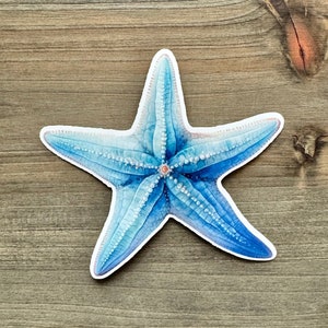 May include: A blue starfish sticker with a white border. The starfish has a detailed watercolor design with varying shades of blue and white. The sticker is placed on a wooden surface.