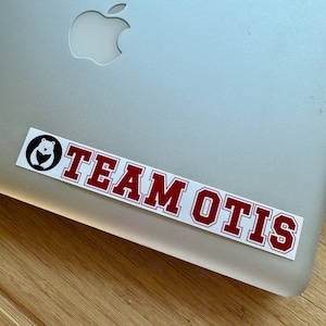 May include: A white sticker with a red border on a silver laptop. The sticker features a black and white illustration of a bear and the text "TEAM OTIS".