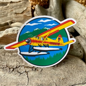 May include: A yellow and red seaplane with white floats, flying over a blue lake with green trees and snow-capped mountains in the background. The plane has the text "N380FG" on the side.