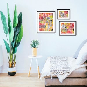 May include: Three framed abstract art prints with black frames and white floral designs on a colorful background. The prints are hanging on a white wall above a gray couch with a white throw blanket.