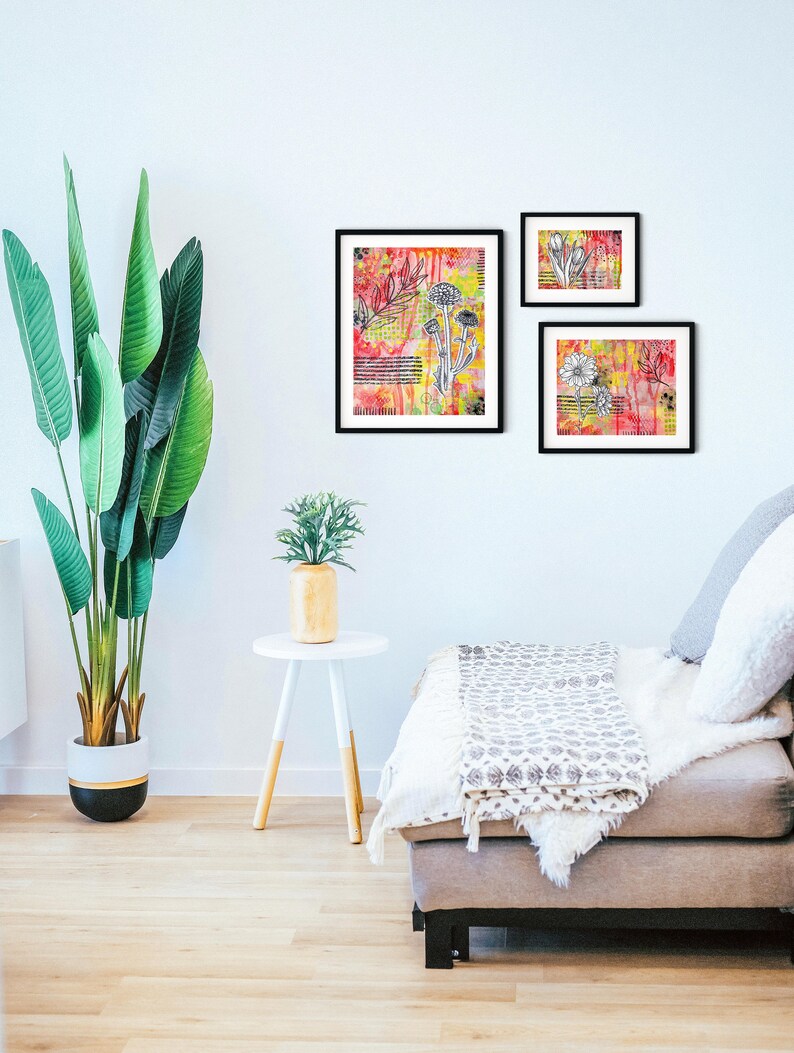 May include: Three framed abstract art prints with black frames and white floral designs on a colorful background. The prints are hanging on a white wall above a gray couch with a white throw blanket.