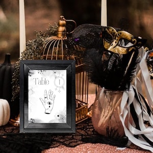 May include: A black framed table number card with a white background and a black and white illustration of a hand with an eye in the palm. The card says "Table 1" in black text.