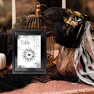 May include: Black framed table number card with the text "Table 1" and a black and white illustration of a sun with an eye in the center. The card is on a table with a black and white tablecloth and a birdcage, candles, and a masquerade mask.