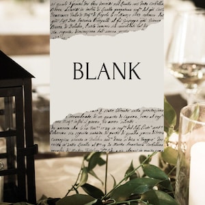May include: A blank, torn-edge card with the word "BLANK" in a serif font. The card has a vintage aesthetic, with handwritten text visible in the background. Greenery and candles are in the foreground.
